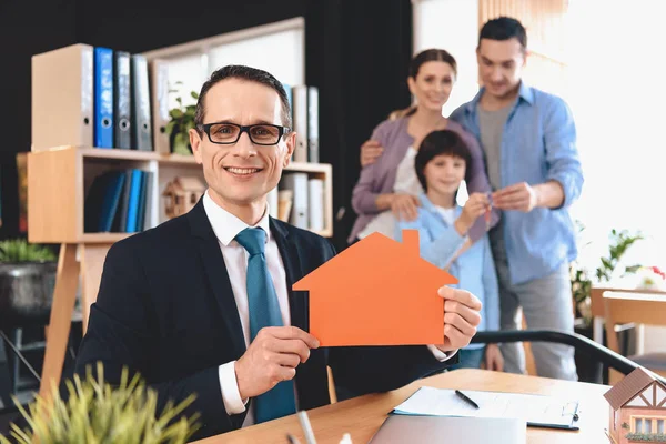 Makler sitzt am Schreibtisch im Büro. Makler präsentiert Haus-Ikone mit Familie im Hintergrund. — Stockfoto