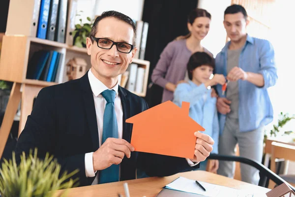 Makler sitzt am Schreibtisch im Büro. Makler präsentiert Haus-Ikone mit Familie im Hintergrund. — Stockfoto