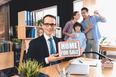 Emlak ofis masasında oturuyor. Emlak ana sayfa Satılık işareti için aile arka planda gösteren.