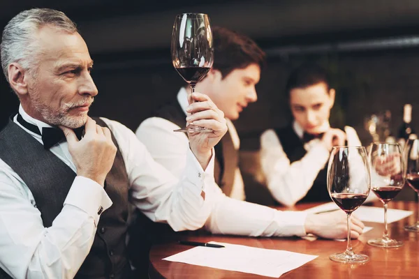 Viejo sommelier experimentado en corbata de lazo está sosteniendo un vaso de vino tinto mientras está sentado en la mesa . — Foto de Stock