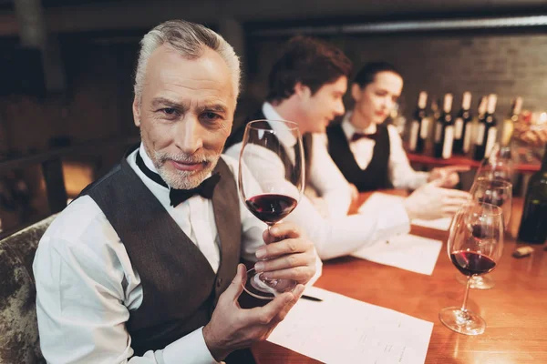穿着领结的老侍酒师坐在桌旁拿着一杯红酒. — 图库照片