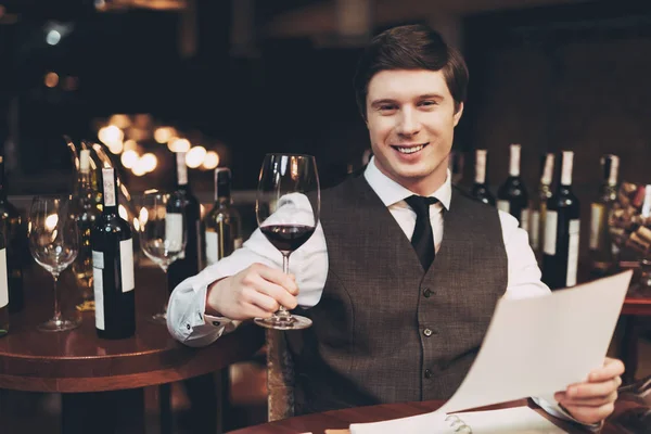 Χαρούμενο, κομψό νεαρός άνδρας που κρατά ποτήρι κόκκινο κρασί και το κρασί λίστα στα χέρια. — Φωτογραφία Αρχείου