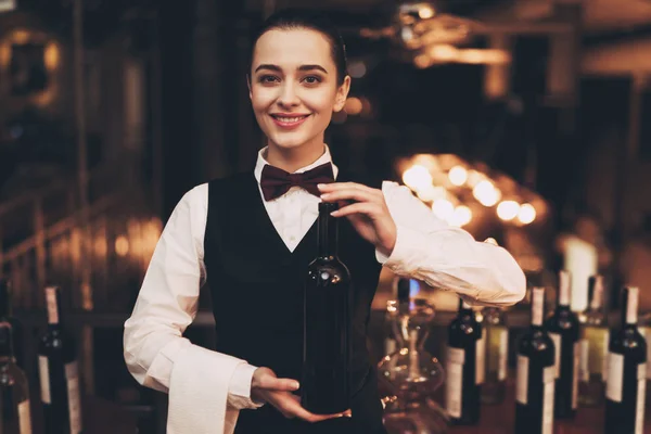 Χαρούμενη σερβιτόρα κομψό κρατώντας το μπουκάλι κόκκινο κρασί, στέκεται κοντά στο μπαρ. — Φωτογραφία Αρχείου