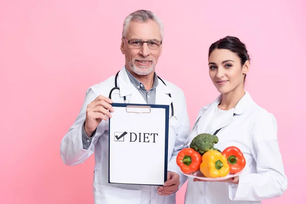Portrait of smiling male and female doctors with stethoscopes holding diet sign and plate of vegetables isolated.