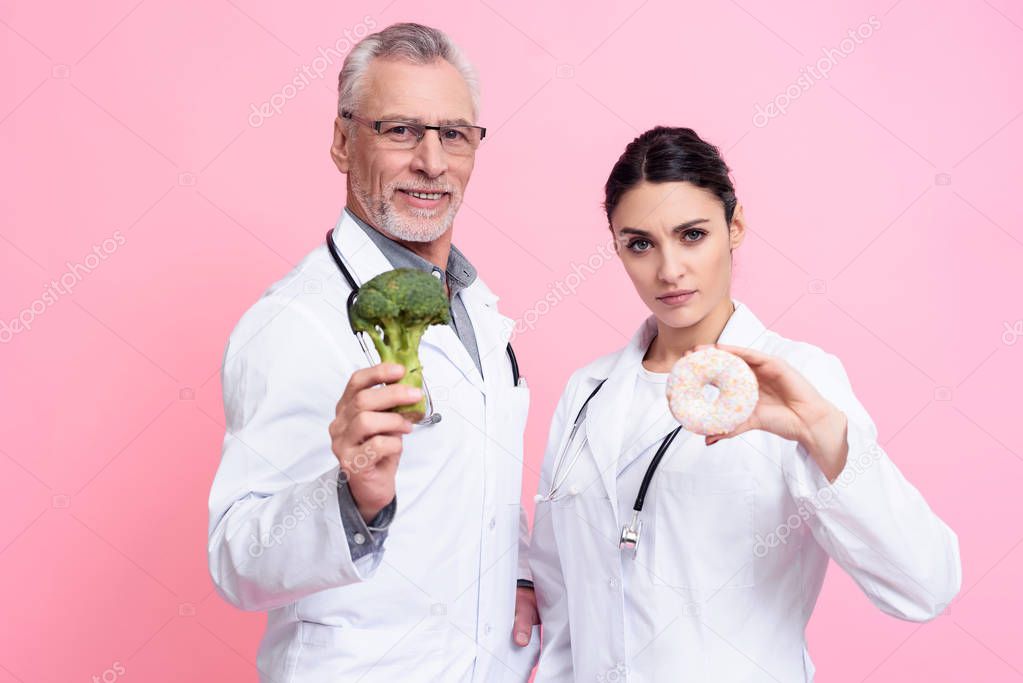 Portrait of male and female doctors with stethoscopes holding broccoli and donut isolated.