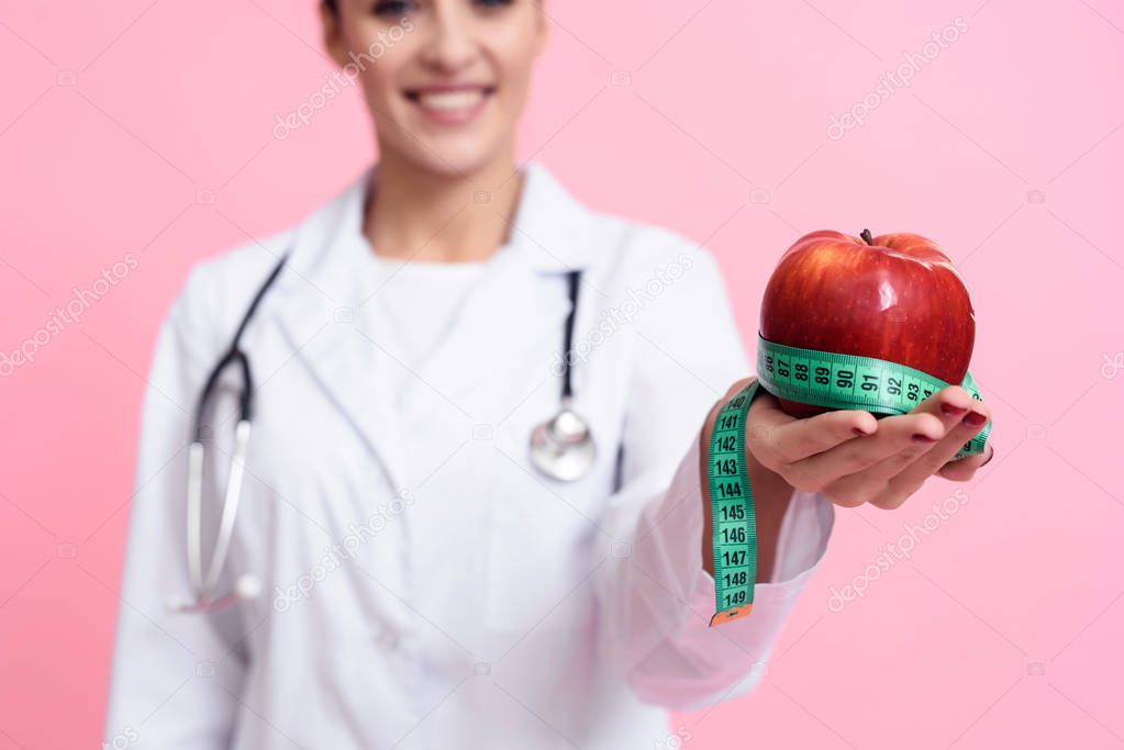 Portrait of smiling female doctor with stethoscope holding measuring tape and apple isolated.