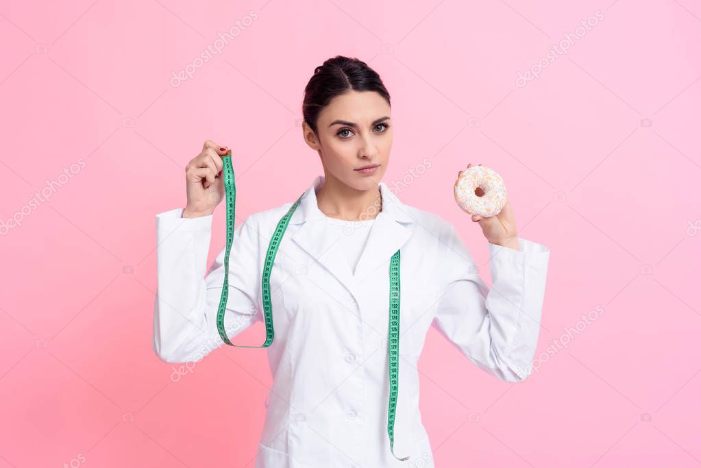 Portrait of female doctor with stethoscope holding measuring tape and donut isolated.
