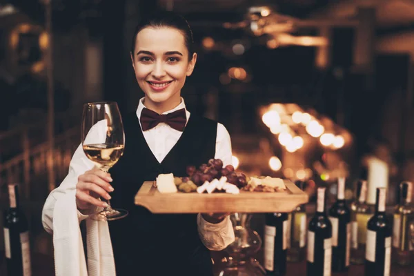 Χαρούμενη σερβιτόρα κομψό κρατώντας το ποτήρι λευκό κρασί, πιάτο τυριού και φρούτων, στέκεται κοντά στο μπαρ. — Φωτογραφία Αρχείου