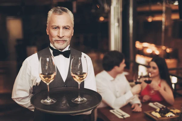 Sommelier está sosteniendo bandeja con copas de vino blanco, de pie cerca de la mesa de la pareja que están en la fecha . — Foto de Stock