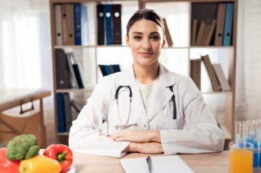 Female doctor sitting at desk in office with clipboard and stethoscope.