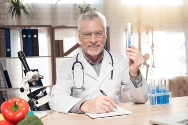 Doktor mikroskop ve stetoskopla ofiste masada oturuyor. Adam elinde ilaç bardağı tutuyor..