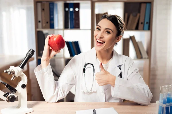 顕微鏡と聴診器オフィスで机に座っている女性医師。女性は赤いリンゴを保持しています。. — ストック写真