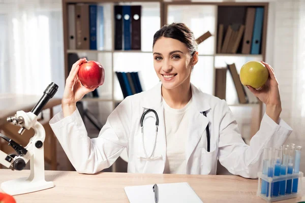 顕微鏡と聴診器オフィスで机に座っている女性医師。女性は黄色と赤のリンゴを保持しています。. — ストック写真