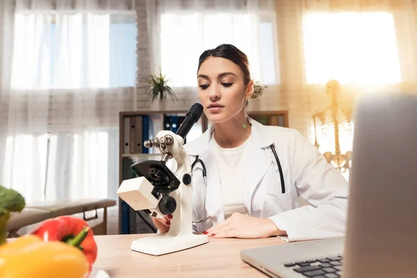 顕微鏡と聴診器オフィスで机に座っている女性医師。女性が顕微鏡で見てください。. — ストック写真