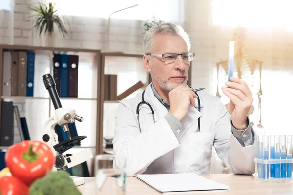 顕微鏡と聴診器でオフィスの机に座っている医師。男は薬のガラスを持っている. — ストック写真