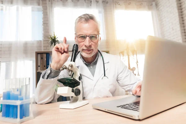 医師は顕微鏡と聴診器のオフィスで机に座って。男性は顕微鏡で見ています。. — ストック写真