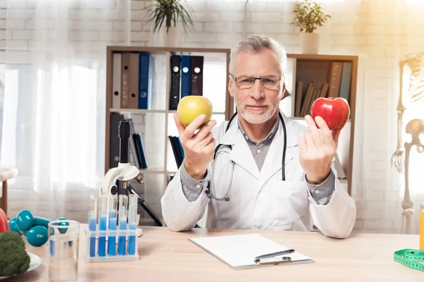 Doctor sitting at desk in office. Doctor is holding fresh red and yellow apples. — ストック写真