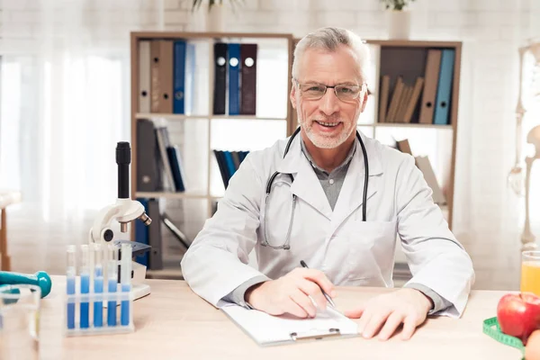 Doctor sitting at desk in office. Man is writting on clipboard. — ストック写真