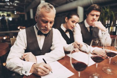 İki erkek sommeliers ve kadın sommelier şarap gözlük ile masada oturan şarap listesi oluşturur.