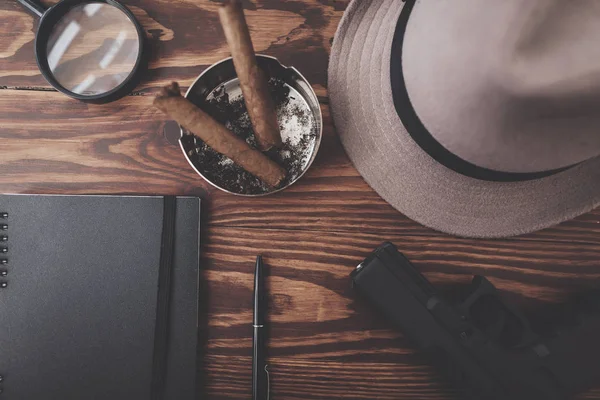 Widok z góry. Szkło powiększające, sigars, kapelusz, pistolet i notebook z pióra na biurko. — Zdjęcie stockowe