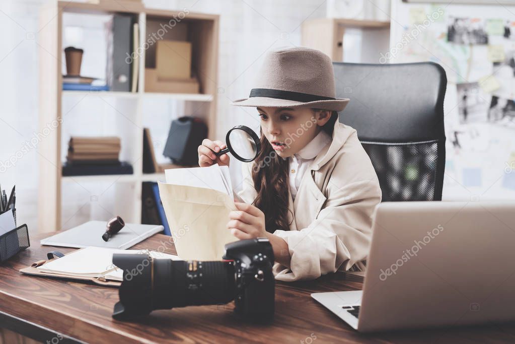 Private detective agency. Little girl is sitting at desk looking at photos with magnifying glass.