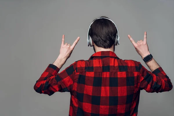 后面的视图 用耳机听音乐的人 年轻的褐发女郎家伙在红色检查衬衫显示手势摇滚标志 在灰色背景上隔离 人的情感观念 — 图库照片