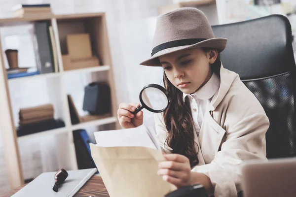 Private detective agency. Little girl is sitting at desk looking at photos with magnifying glass. — Stock Photo, Image