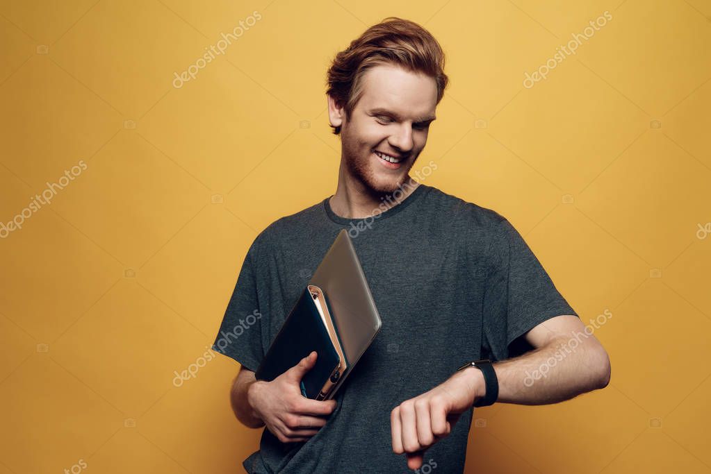 Portrait of Cheerful Young Guy Holding Laptop. Man Looks on Watch Smiling Laughing. Time managment and productive daylife. Isolated on Orange Background. Concept of Humans Emotions