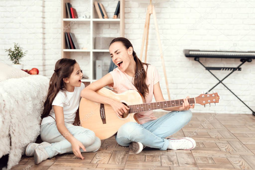 Mom and her daughter are sitting on the floor at home and playing the guitar. They sing to the guitar.