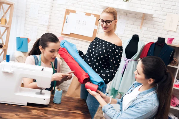 Three girls at garment factory. They are sitting behind sewing machines and choosing fabric for new dress. — Stock Photo, Image