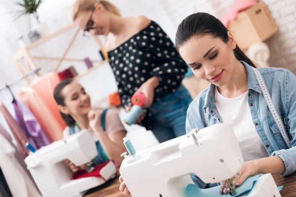 Three girls at garment factory. They are sitting behind sewing machines and choosing threads for new dress. — Stock Photo, Image