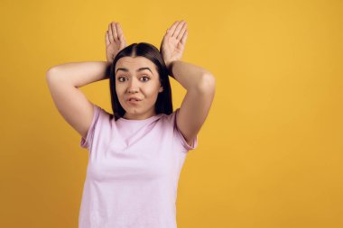 Genç kız tavşan, kafası gibi kulakların arkasında ellerini koyarak gösterir. Sarı arka plan üzerinde izole. Stüdyo portre.