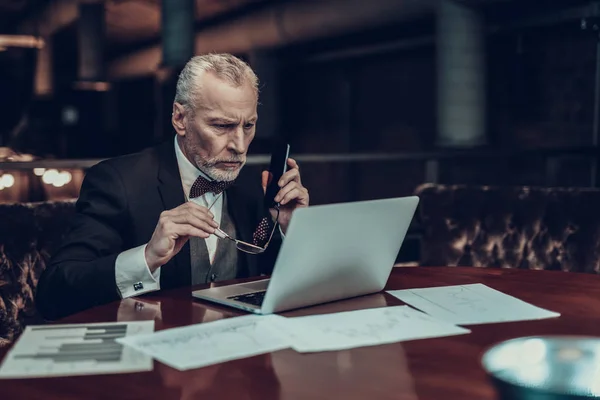 Businessman Using Laptop. Old Businessman . Man is Wearing in Black Suit. Experienced Entrepreneur. Strict Man. looking at Laptop. holding Pnone and glasses . Working Late. concentrated Person
