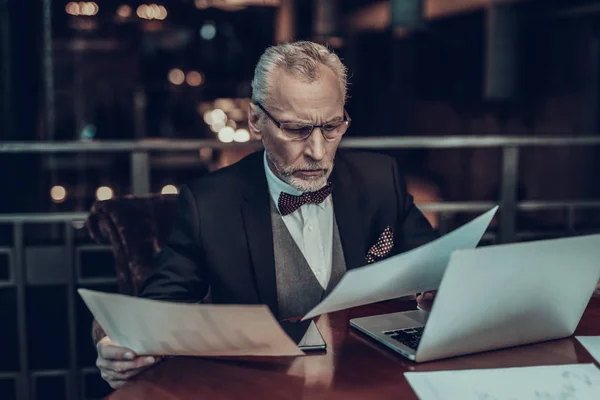 Businessman Using Laptop. Old Businessman . Man is Wearing in Black Suit. Experienced Entrepreneur. Strict Man. looking at Graphics. holding piece of paper. Working Late. concentrated Person