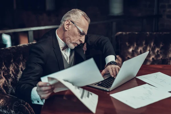 Businessman Using Laptop. Old Businessman . Man is Wearing in Black Suit. Experienced Entrepreneur. Strict Man. looking at Laptop. Working Late. holding piece of paper. talking on Phone .