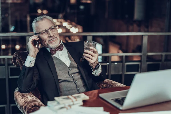 Businessman Using Laptop. Old Businessman . Man is Wearing in Black Suit. Experienced Entrepreneur. Strict Man. looking at side. talking on Phone. stack of money on table. holding a glass