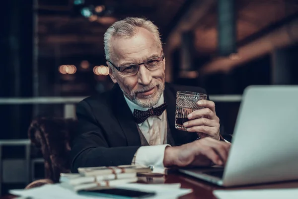 Businessman Using Laptop. Old Businessman . Man is Wearing in Black Suit. Experienced Entrepreneur. Strict Man. stack of money on table. holding a glass of cognac. portrait of old man
