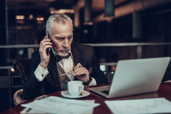 Businessman Using Laptop. Old Businessman . Man is Wearing in Black Suit. Experienced Entrepreneur. Strict Man. looking at Laptop. talking on Phone. glasses near mouth. cup of coffee on table
