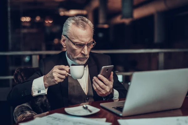 Businessman Using Laptop. Old Businessman . Man is Wearing in Black Suit. Experienced Entrepreneur. Strict Man. looking at Phone. talking on Phone. holding cup of coffee. Laptop on table.
