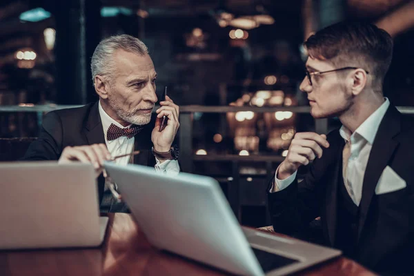 Businessman Using Laptop. Old Businessman . Man is Wearing in Black Suit. Experienced Entrepreneur. Strict Man. Phone near face. Laptop on table. conversation. look at each other. Business Meeting