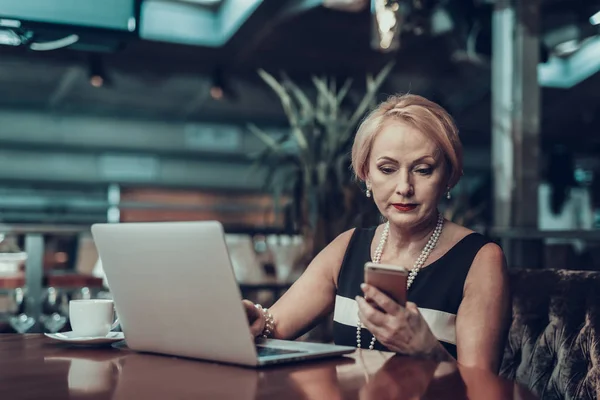 Businesswoman Using Laptop. Old Businesswoman looking on Phone. Experienced Entrepreneur. Strict Woman. Working Late. Laptop on table. Success Grandmother. Sits in Office. Solves Problems.