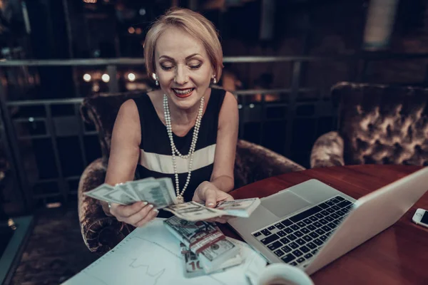 Businesswoman Using Laptop. Old Businesswoman looking Laptop . Experienced Entrepreneur. Working Late. Success Grandmother. Sits in Office. smiling woman. holds and counts money. Graphics on table