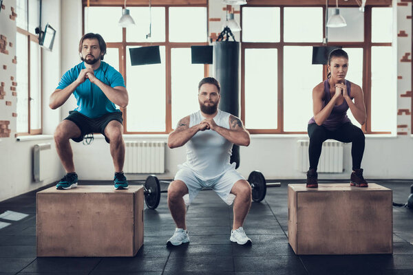 Two Men And Woman Doing Squats Exercises. Training Day. Fitness Club. Healthy Lifestyle. Powerful Athlete. Active Holidays. Crossfit Concept. Spacious Bright Gym. Comfortable Sportswear.