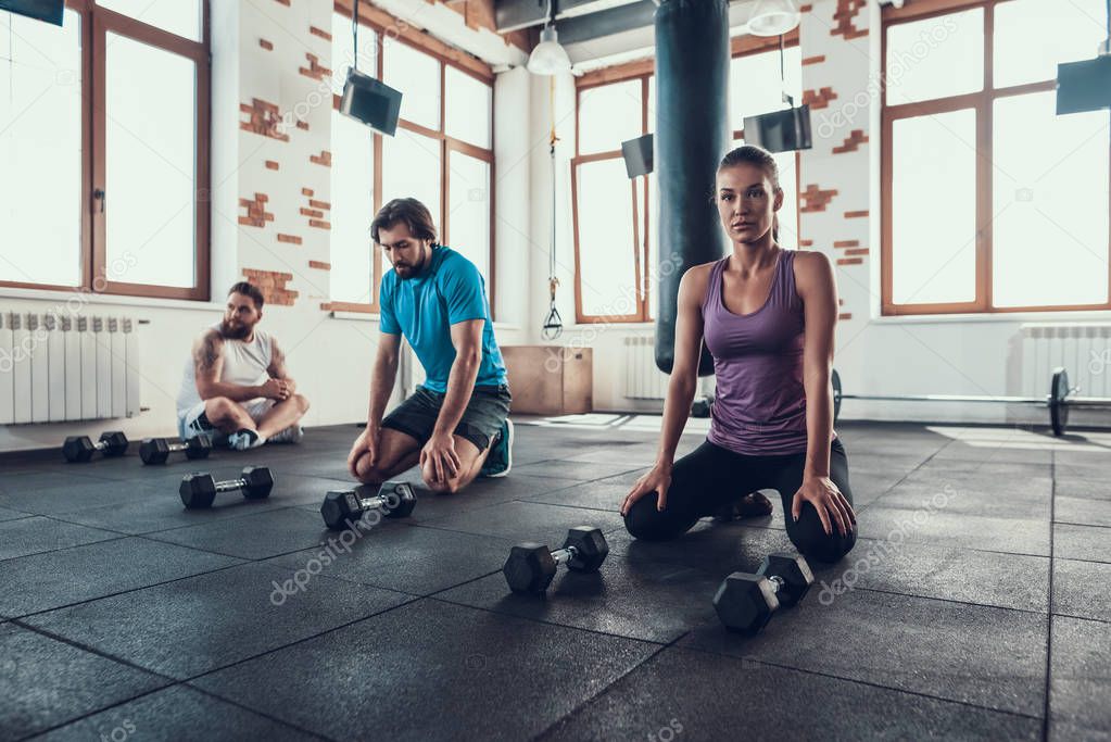 Two Guys And Girl Rest On Gym Floor. Break Time. Training Day. Fitness Club. Healthy Lifestyle. Active Holidays. Crossfit Concept. Tired Athletes. Sit With Dumbbells. Depleted Sportsmen.