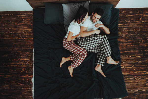 Couple is Lying on Bed. Couple is Young Beautiful Woman and Man. Woman is Hugging Man. Persons is Happy and Smiling. People is Wearing Pajama Pants and T-Shirts. Home Interior. Top View.