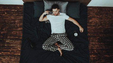 Lying yatak yalnız üzerinde bir adamdır. Genç güzel beyaz adam giyen pijama pantolon ve T-Shirt kişidir. Elinde tabanca başında bir adamdır. Çalar saat Lying yatak var. Ev iç yüz yüze. Üstten görünüm.