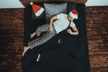 Lying yatak yalnız üzerinde bir adamdır. Genç güzel beyaz adam Noel şapka giyen kişidir. Adam içki şampanya şişe var. Kişi iç ana sayfa bulunur. Adamın üstten görünüm.