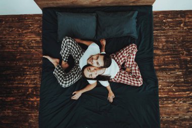 Çift yatağa sırt sırta oturuyor. Çift genç güzel kadın ve erkek kişi seyir yukarıya ve gülen. Pijama pantolon giyen ve t-shirtler kişidir. İnsanlar ev iç. Üstten görünüm.