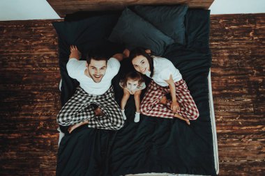Yatakta mutlu bir aile. Anne, baba ve kızı ailedir. Kişi arıyorum kalmış. Ebeveynler Smiling olduğunu. Pijama pantolon giyen ve t-shirtler kişidir. İnsanlar ev iç. Üstten görünüm.