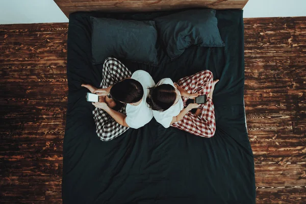 Couple is Sitting on Bed Back to Back. Couple is Young Beautiful Woman and Man. Persons is Using Mobile Phones. People is Wearing Pajama Pants and T-Shirts. People in Home Interior. Top View.